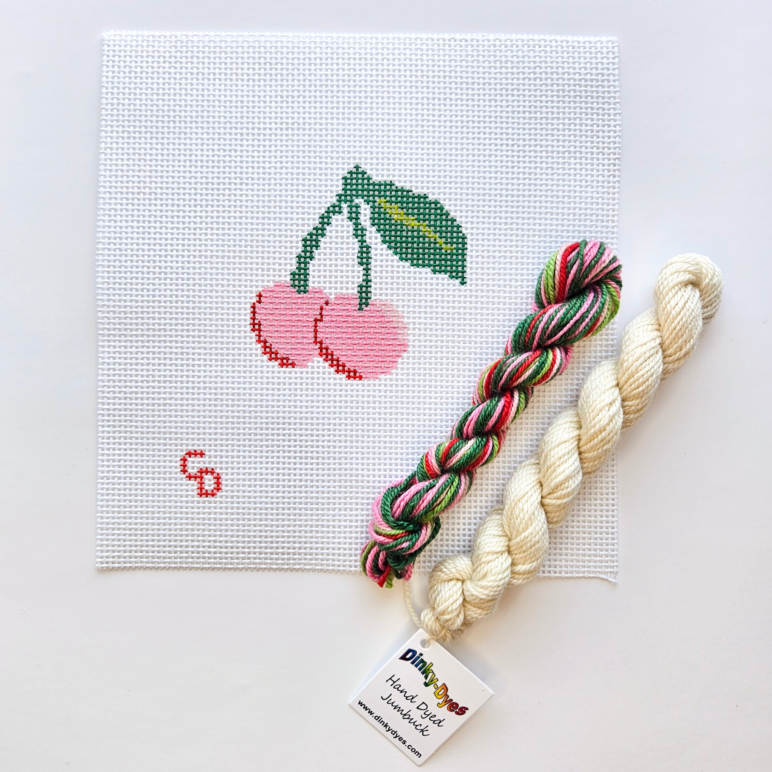 Needlepoint kit with cherry design and two skeins of yarn on a white background