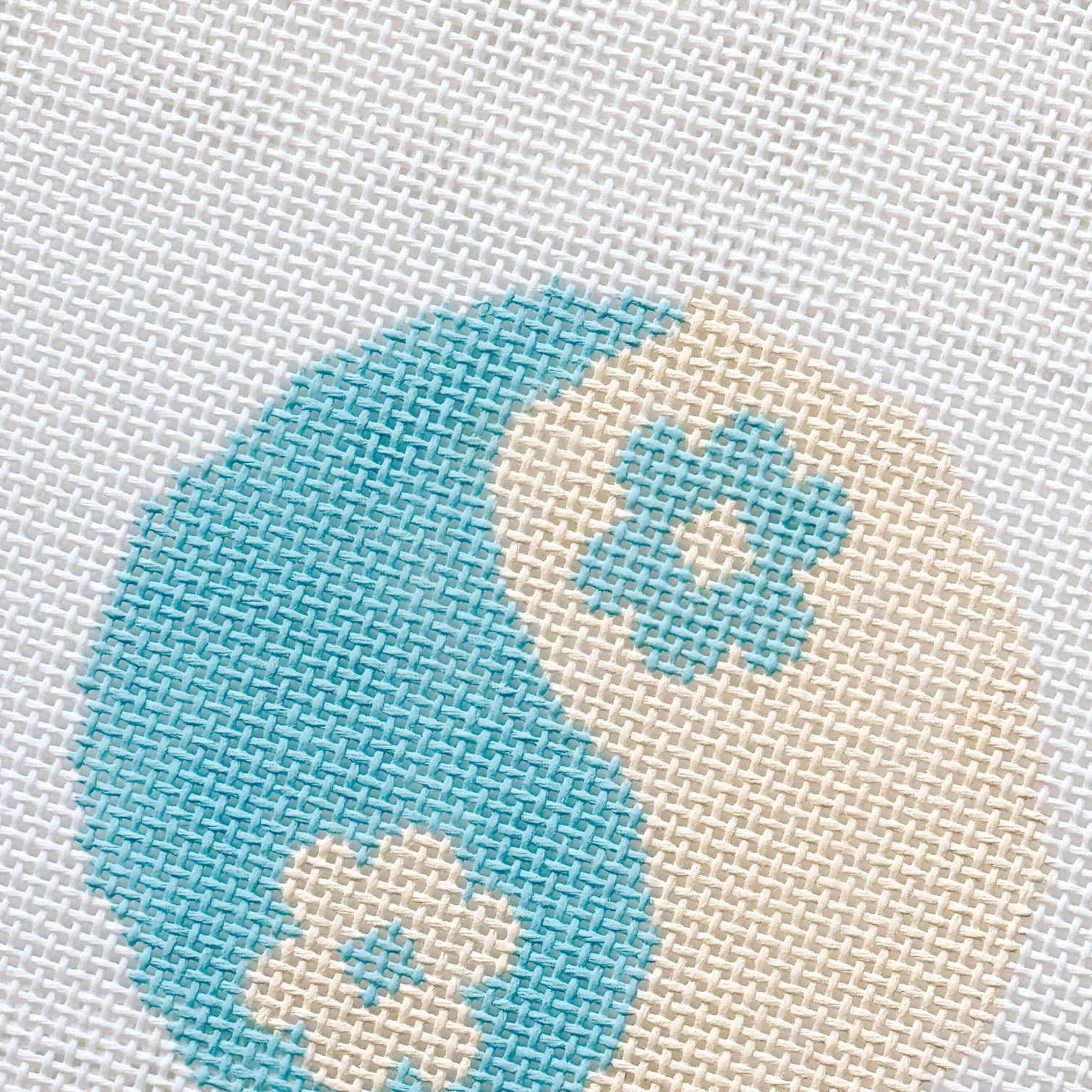 Needlepoint design of a yin yang symbol with blue and beige colors on a textured fabric background.

