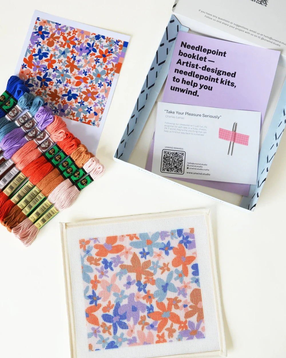 Needlepoint kit with floral patterns, threads, and a booklet on a white surface.